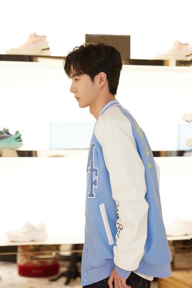 A side view of Wang Yibo at an event to promote the opening of a new Moncler store in Shanghai. He wears a pale blue varsity style jacket with the letters MF on one side. He stands in front of brightly backlit shelves displaying athletic shoes.