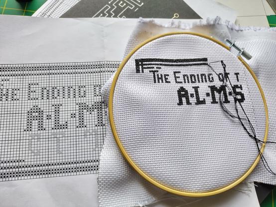 A partly finished cross-stitch sitting on the paper pattern; the only words visible are "The ending of t—" and "ALMS—"