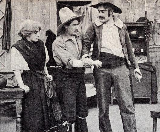 A still of the film. We can see two men and a woman. The woman has tied hair. She is wearing a long, dark skirt and a short-sleeved white blouse. The men are wearing big cowboy hats, dark trousers and shirts.