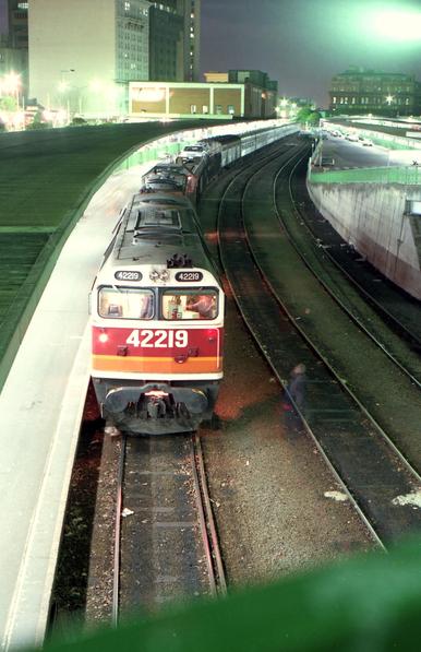 A night time long exposure image taken from an overbridge looking down onto a loco hauled passenger train standing at a curved platform at a city terminal station.  The train, which is of stainless steel fluted body carriages with a motorail car carrying wagon, is headed by two diesel locomotives - the leading one of which is a 422 class boxcab style unit painted in NSW SRA Candy livery.  The ghostly image of a railwayman in overalls is captured standing on the adjacent track looking up at the loco cab talking to the train driver.