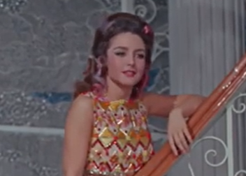 Angelica María posando en una escalera, vistiendo un vestido colorido con un diseño geométrico, con cabello peinado y un ambiente vibrante en el fondo.