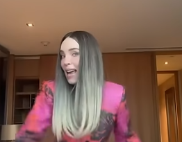 Belinda, vistiendo un blazer rosa con negro, sonríe y gesticula en un entorno interior bien iluminado.