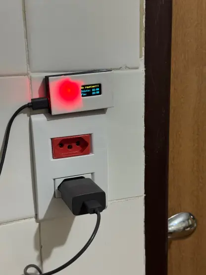 Uma parede e se vê uma porta do lado direito. Na parede tem uma tomada que é o foco da imagem. Nela tem um carregador USB ligado e o fio vai até uma caixinha que tem a frente branca. A caixa tem uma luz vermelha forte e uma pequena telinha e na tela se lê "EM REUNIÃO" e logo abaixo desse texto tem os horários de início e fim da reunião.
