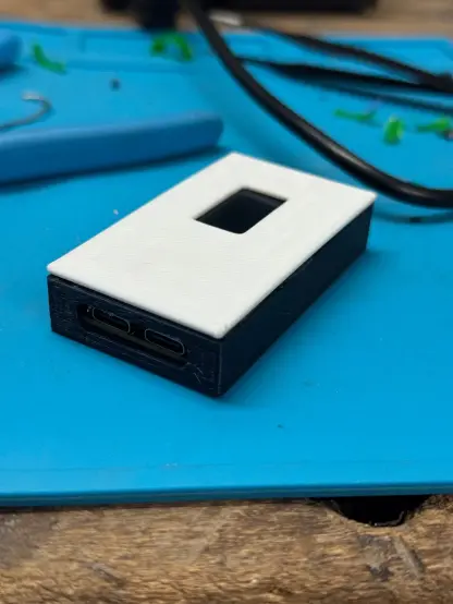 Visão isométrica da caixinha. Se vê plugs USB em uma face, e a tela OLED na superfície
