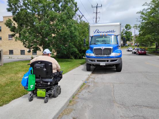 A man on a motorized wheelchair  is facing a large truck parked so that it blocks half of the sidewalk.