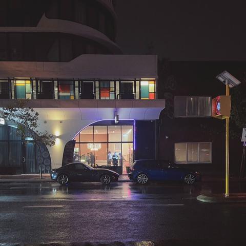 Diners enjoy a desert bar on a cold rainy night. Two cars are parked on the street in front of the desert shop. Multicoloured windows appear above it. To the right is a LED sign saying 40.