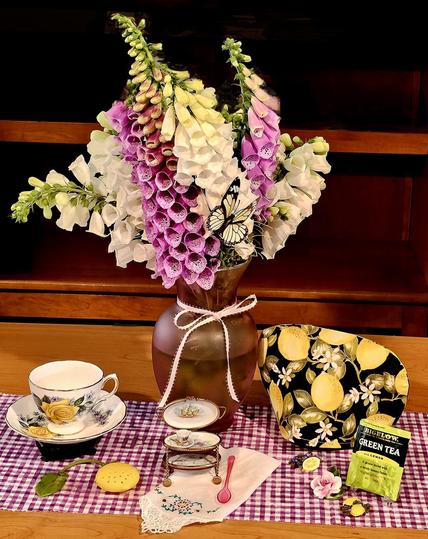 Tea tableau in purple and yellow with a vase of foxgloves and lemon- themed cozy and knickknacks, vintage china, and a vintage French trinket box that is a miniature functioning tea cart. The tea is Bigelow green tea with lemon.