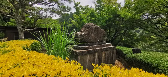 A stone monument in a Japanese garden with a large, rough-hewn stone on a tiered base surrounded by lush greenery, yellow flowers, and trees, conveying tranquility and natural beauty.