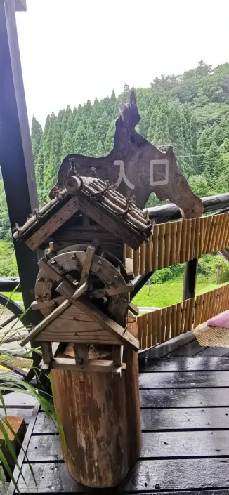 A wooden water wheel with a small roof mounted on a post, placed in front of a sign reading "入口" (entrance), surrounded by a lush green forest.