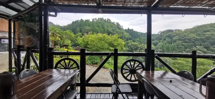 A wooden deck with two dark tables and chairs, offering a view of green hills, trees, and a winding paved road. The bamboo roof and wagon wheel rail decorations are visible.