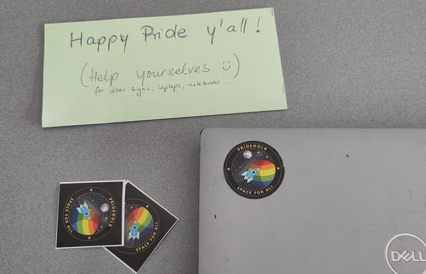 A number of stickers laying on a table, one is put on my work laptop. The stickers show planets in different pride colours and a smal spacecraft. They have a text reading "pride @ DLR - space for all".

Next to it: A handwritten sign "Happy Pride y'all (Help yourselves) for door signs, laptops, notebooks ...
