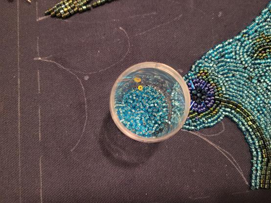 A container of seed beads from above. The project they're being used in is to the right. The beads are sky blue, but there is one bright yello,w slightly larger, bead mixed in by mistake.