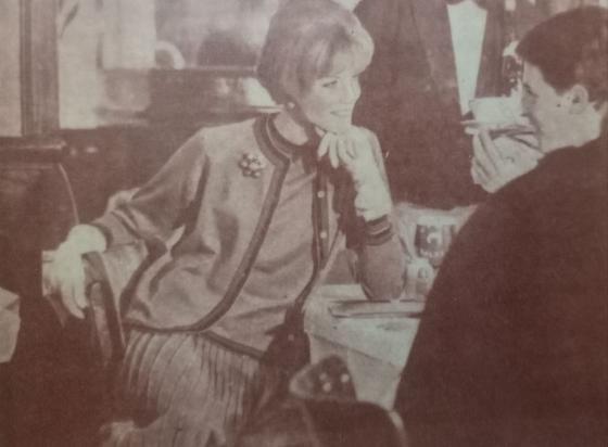 Una mujer sonriente en un café, mirando con atención a un hombre que le habla, mientras en la mesa hay una bebida y otros clientes de fondo.