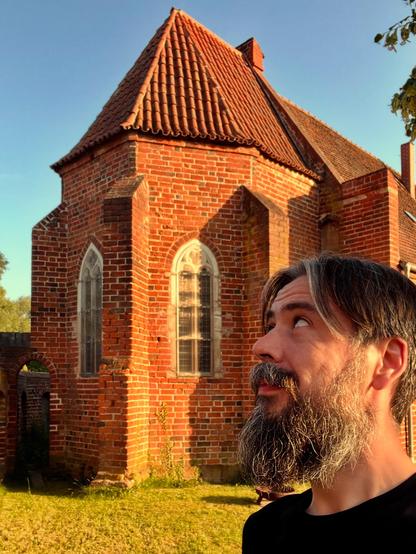 Ein Mann mit Bart blickt auf ein historisches Backsteingebäude mit einem markanten Spitzdach. Die Szene wird von warmem Sonnenlicht beleuchtet, und die Umgebung ist von grünem Gras und Bäumen geprägt.