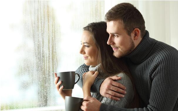Happy couple in thick, gray turtleneck sweaters sipping on gray mugs looking out a rainy window