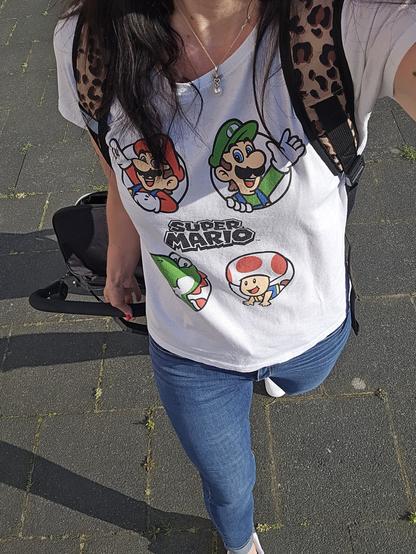 Das Bild zeigt eine Person, die ein weißes T-Shirt mit dem "Super Mario"-Logo trägt. Auf dem T-Shirt sind die Charaktere Mario, Luigi, Yoshi und Toad abgebildet. Die Person trägt eine Jeans und einen Rucksack mit einem Leopardmuster. Sie steht auf einem gepflasterten Weg, und im Hintergrund ist ein Teil eines schwarzen Gepäcksstücks zu sehen. Die Person hat langes, dunkles Haar und trägt eine Halskette mit einem Perlenanhänger.