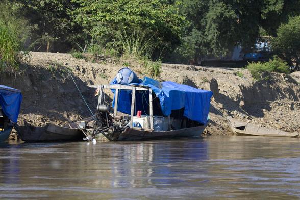 Blick von der Flußseite auf ein lehmiges Steilufer. Im Wasser vor dem Ufer liegen Holzboote. Die beiden links und rechts zu sehenden Boote haben keinerlei Aufbauten oder Antrieb. Nur das Boot in der Mitte ist mit einer über ein Holzgestell gezogenen blauen Plane überdacht. An Bord stehen Kühltruhen. Am Heck befindet sich ein Benzinmotor, der den Propeller antreibt. Die Konstruktion wirkt wie selbst gebaut.
Oberhalb der Uferböschung grüne Büsche und Bäume.