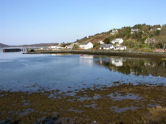 Gairloch Harbour, at Charlestown in Wester Ross. The image shows a view over a bay at low tide to a settlement of white buildings climbing a hillside from the far shore. At its left-hand end is a harbour. The scene is in sunlight.