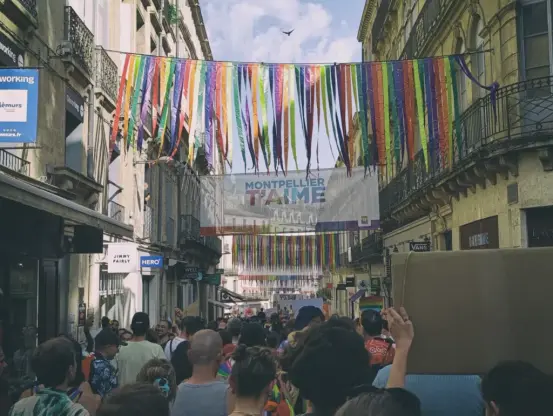 Photo d'une rue commerçante étroite à Montpellier remplie de personnes manifestant pour la Marche des fiertés, décorée de longues bandes de tissu arc-en-ciel suspendues et d’une banderole indiquant "Montpellier t’aime".