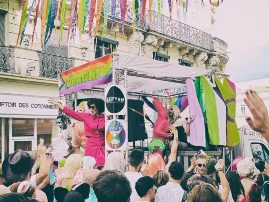 Photo d'un char de la marche des fiertés avec plusieurs drag queens en spectacle sur une estrade décorée de drapeaux LGBTQ+, entourée par une foule dans une rue commerçante de centre-ville.