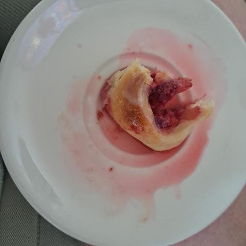 A half eaten sourdough roll sits on a white saucer. The roll is surrounded by a puddle of red, the juices from the fruits. To the left of the photo is my khaki shorts, to the right my legs.