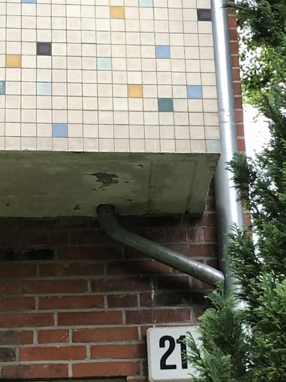 Abschnitt eines Balkons mit Fliesen-Mosaik. Es ist weiß mit vereinzelt eingefügten farbigen Kacheln, ockergelb - hellblau, türkis, antrazit. Darunter Backsteinwand mit Hausnummer 21. Ein Fallrohr verläuft aus dem Balkonboden zum Hauptrohr an der Hausecke. Rechts ein Stück Grünpflanze.