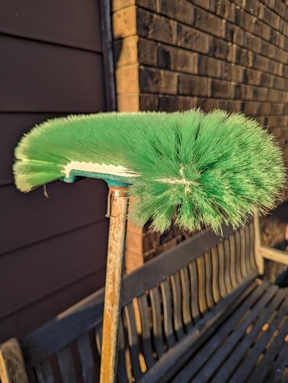 A 60+yo broom basks in winter solstice golden hour sunlight. It has a green nylon head, and the original well worn white broomstick. The nail came later. It's leaning on a wooden bench in front of brown brick and fibro.
