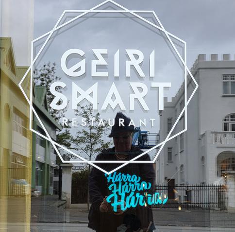 A photograph of a shop window in Reykjavik with latin ALLCAPS lettering bass on runic style of letter forms (straight lines), but keeping a curve kept for the V and the R "GEIRI SMART'. In the background we see a blue, backlit text in an upright cursive  "Hurra"