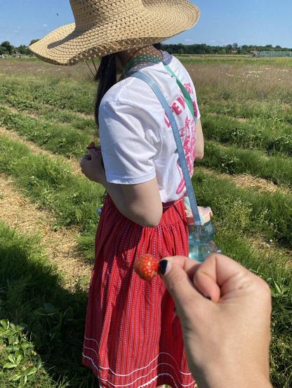 A tiny strawberry is held up by a hand in front of a woman standing in a field with a red patterned skirt, white T-shirt and sunhat on, blue bear waterbottle is  hung by strap over her shoulder