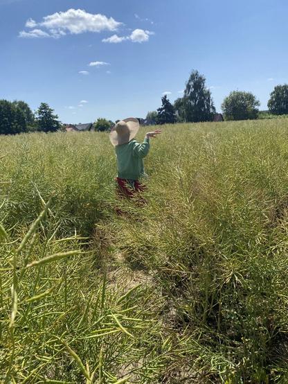 A woman dancing in a field, seen from the back  with a red patterned skirt, green jacket and sunhat on, blue bear waterbottle is  hung by strap over her shoulder, she bends her arm , fingers fanned