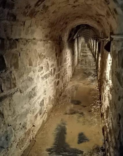 A dimly lit stone tunnel with a curved ceiling and wet, muddy floor. The walls are made of unevenly cut stones, and the tunnel stretches far into the distance with small lights mounted along the right side, casting shadows and reflections in the water.