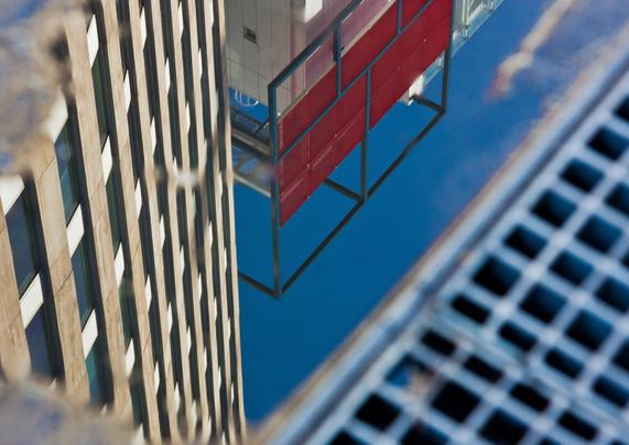 In the right corner, from bottom center to top right, an iron drain grate with small squares. On the left, from bottom center, diagonally upwards, is a beige office building, reflected in a puddle next to the grate with rectangular windows. At the top, a triangle of a building with a black and red steel square decoration. In between, a clear blue sky.