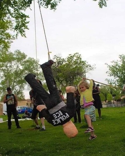 At a birthday party, a young white girl with blond hair in her 6 is holding a long stick with her 2 hands a large smile on her face ready to hit a special pinata hanging from a tree made of a human size doll wearing police garments and written on it's back in huge white letters POLICE ICE