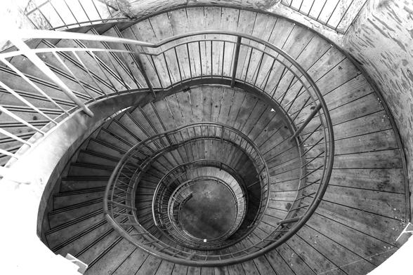 A high-angle, black and white shot looking down a tightly spiralling staircase. The wooden treads form concentric circles, appearing smaller as they descend into the darkness at the bottom. A dark metal handrail snakes around the inner edge of the stairs, while a lighter-coloured, more modern handrail is visible on the outer edge in the top left. The walls enclosing the staircase are light-coloured and textured, possibly stone or concrete.
