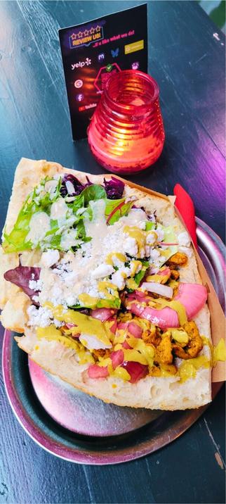 A top-down shot of a vegan döner on a metal plate, featuring a large flatbread folded over a colorful filling. One half of the filling includes green lettuce, purple cabbage, and a white sauce, while the other half contains a yellowish, textured protein, red onions, and a yellow sauce. In the background, on a dark wooden table, there's a small black sign with "REVIEW US!" and social media logos, next to a glowing red lantern.