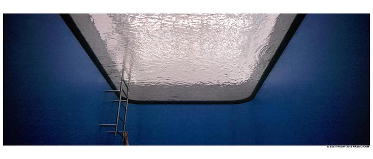 Colour panorama photograph in an art installation. The perspective is as if you are at the bottom of a swimming pool looking up at the surface of the water. The sides of the pool are blue and in the left corner is a metal pool ladder exiting the water. At the bottom of the ladder one arm can be seen hanging on to the ladder. It is an art installation by Leandro Erlich (1973), called Swimming Pool