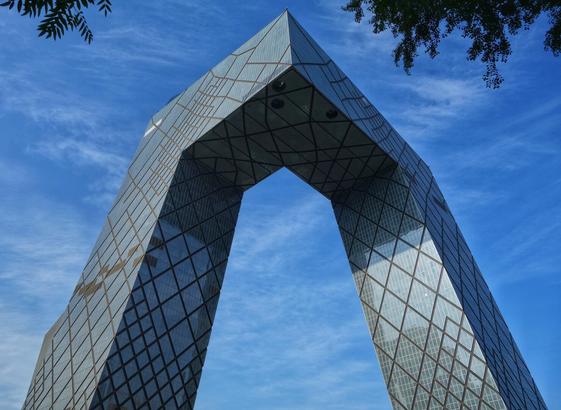 A worm's-eye view captures the unique, angular architecture of the CCTV Headquarters in Beijing against a vibrant blue sky with wispy white clouds. The building, nicknamed "The Big Trousers" or "The Pants" due to its distinctive shape, leans inward, forming a large, trapezoidal archway in its upper section. Its facade is covered in a grid pattern of glass panels, reflecting the sky and surrounding light. Dark green tree branches with leaves are visible at the very top of the frame, one on either side, framing the building.