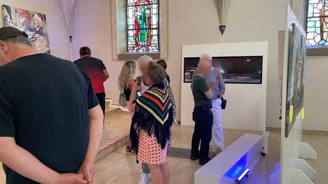 The interior of a profaned church. A bright, vaulted room with large windows. Pictures and photographs hang on the walls and sculptures stand in front of them. The room is filled with people regarding the exhibits