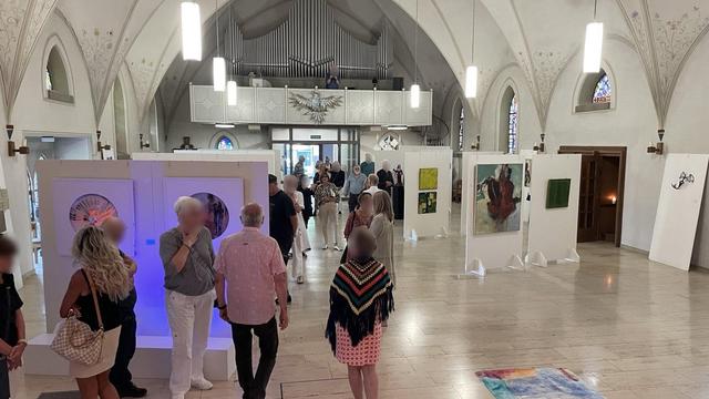 The interior of a profaned church. A bright, vaulted room with large windows. Pictures and photographs hang on the walls and sculptures stand in front of them. The room is filled with people regarding the exhibits