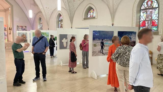 The interior of a profaned church. A bright, vaulted room with large windows. Pictures and photographs hang on the walls and sculptures stand in front of them. The room is filled with people regarding the exhibits