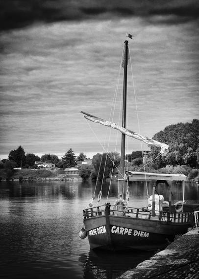 Ein Schwarz-Weiß-Bild eines Bootes mit dem Namen "Carpe Diem", das an einem Flussufer angedockt ist. Das Boot hat einen hohen Mast und ein Segel, im Hintergrund sind Bäume und Häuser zu sehen, die sich im ruhigen Wasser spiegeln.