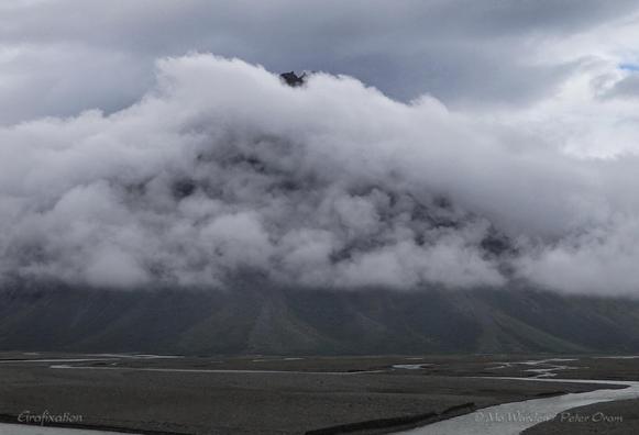 A photo of a mountain, in colour but looking very monochrome. The foreground is flat dark brown sandbanks, cut with channels of flowing water. The foot of the mountain is visible but its flanks are draped with billows of cloud, lit from above. The black peak of the mountain is showing above the clouds.
