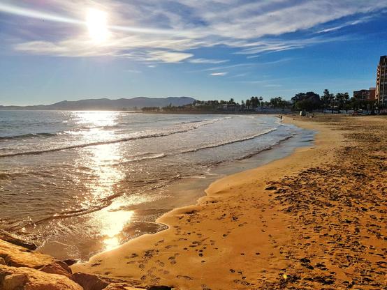 A wide-angle shot of a sunny beach at golden hour, with the sun low in the sky creating a bright glare over the sparkling ocean. Gentle waves roll onto the golden sand, which shows numerous footprints. To the left, dark rocks line the shore, and in the distance, hills and a town with buildings are visible under a blue sky with scattered clouds.