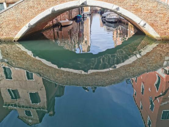 Spiegelung in Venedig. Eine Brücke spiegelt sich im Kanal. Nur in der Spiegelung sind zwei venezianische Häuser und zwei Personen auf der Brücke zu sehen. Unter der Brücke sind im Hintergrund geparkte Boote.