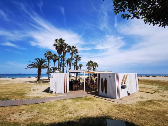 Das Bild zeigt das Strandlokal Km0 in Cambrils, dahinter Palmen und blauer Himmel