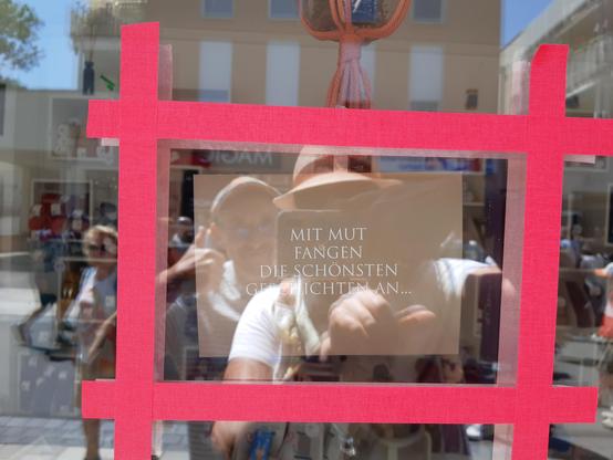 Eine Schaufensterscheibe, darin ist von innen ein Rahmen aus Rosa Klebestreifen geklebt. Darin hängt eine hellbraune Karte, auf der mit weißen Buchstaben steht: MIT MUT FANGEN DIE SCHÖNSTEN GESCHICHTEN AN. In der Scheibe spiegeln sich die Fotografin und ihr Mann, die beide den Daumen nach oben recken.