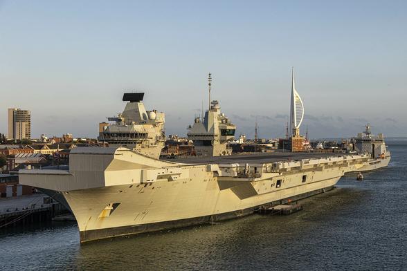 It's early evening on a clear day and we're cruising out of a city's port, passing by the naval dockyard where the main view is that of an aircraft carrier. The vessel has a jump ramp on its port side forward which is nearest to us, and the overall standard grey paint is glowing almost golden with the light of the setting sun hitting it side-on. Behind the aircraft carrier is the sprawling dockyard area and in the distance to the left there's a tower block while the distinctive shape of a tall tower in the shape of a skeletal spinnaker sail marks the exit to the harbour far back to the right.
