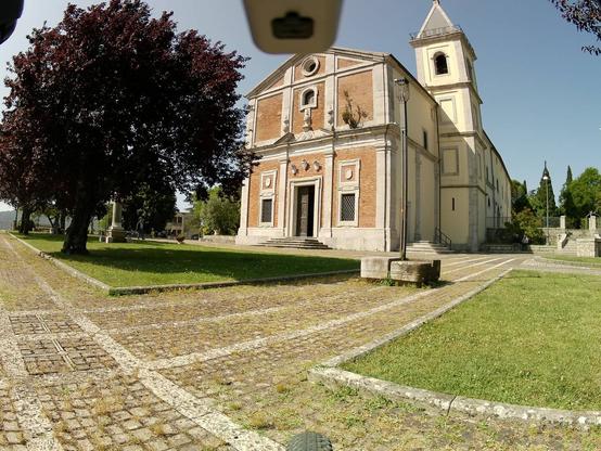 Chiesa in un piazzale con prati, alberi e alcuni manufatti in pietra, tra cui un pozzo, delle scale e una colonna che sorregge un busto, occultato da un albero. Nella foto, visibili anche degli elementi di una bicicletta, tra i quali il più riconoscibile è l'accenno della ruota anteriore.