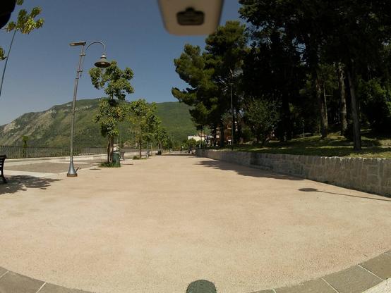 Viale con panchine sulla sinistra, di fronte al belvedere. A destra, gli alberi di una zona verde e in sfondo le montagne. Visibili alcuni elementi di una bicicletta, il più riconoscibile è la ruota anteriore.