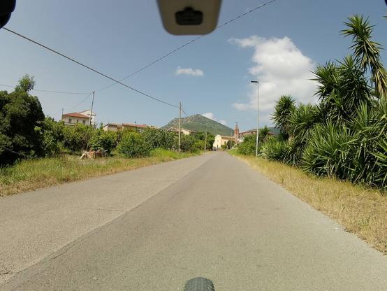 Stradina tranquilla di campagna, con una breve rampa che conduce a una chiesetta, visibile in sfondo.
Visibili alcuni elementi di una bicicletta, il più riconoscibile è la ruota anteriore.